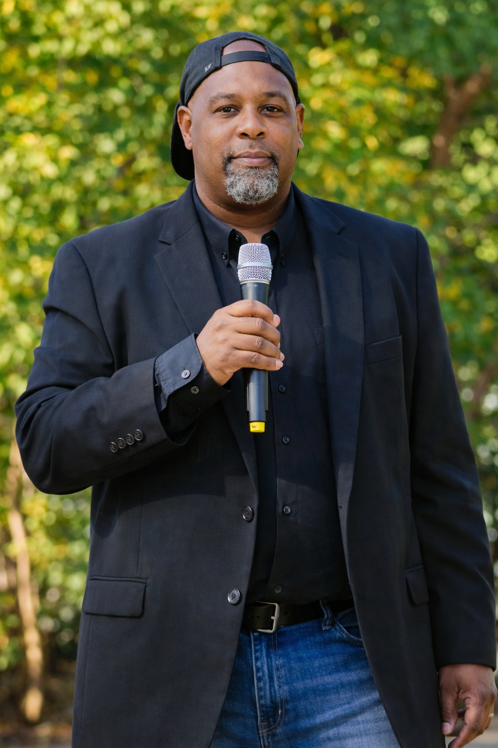 Man in a black blazer and jeans holds a microphone outdoors, wearing a backward baseball cap.