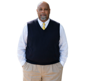 Man standing in a portrait pose wearing a white shirt, navy sweater vest, yellow tie, and beige pants in a business-casual outfit