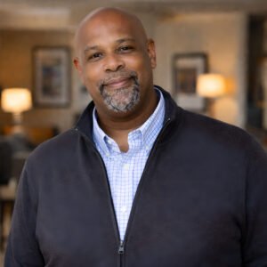 Smiling African American man with a gray goatee wearing a blue checkered shirt and dark zip-up jacket indoors under warm lighting.
