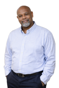 Smiling man in a light blue checkered shirt with hands in his pockets.