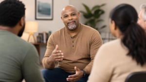 Man in a brown long-sleeve shirt speaks during a group discussion in a cozy living room setting, others listening nearby.