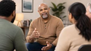A man with a shaved head and goatee speaks, gesturing with his hands during a group discussion in a cozy room.
