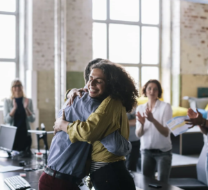 Two coworkers hug in an office while teammates applaud in the background.
