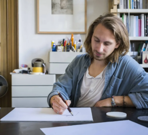 Young man with shoulder-length hair in a denim shirt sketches on white paper at a cluttered art studio desk with supplies in the background.