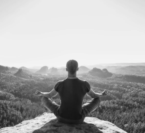 Person seated cross-legged on a rocky cliff, meditating over a vast forest valley seen in black and white.